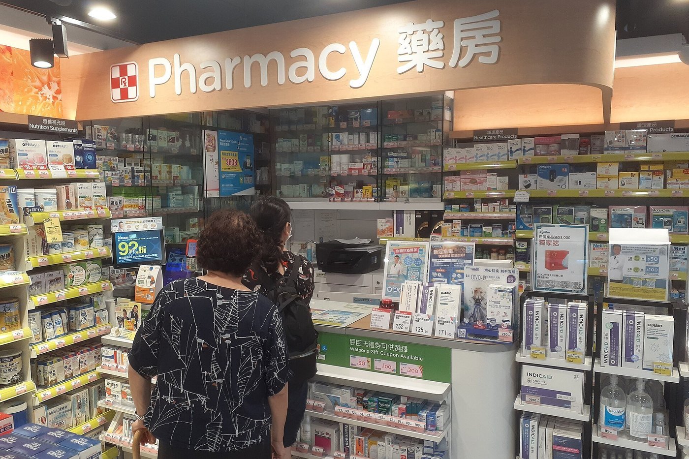 Een klant bij de balie van een apotheek met volle schappen medicijnen.