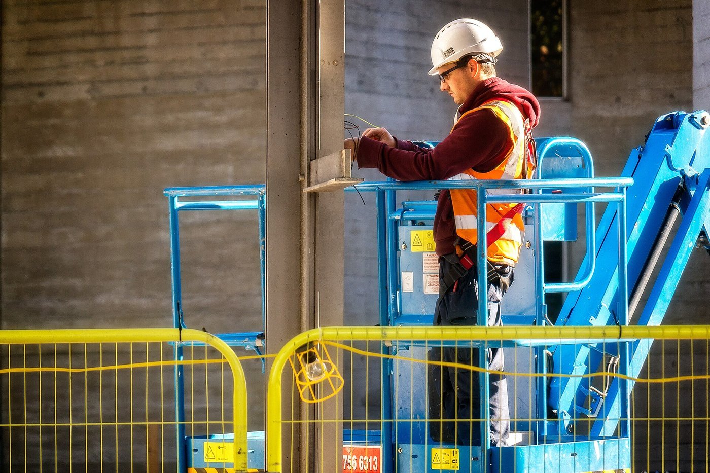 Een monteur met helm werkt op een hoogwerker aan een installatie in een hal.