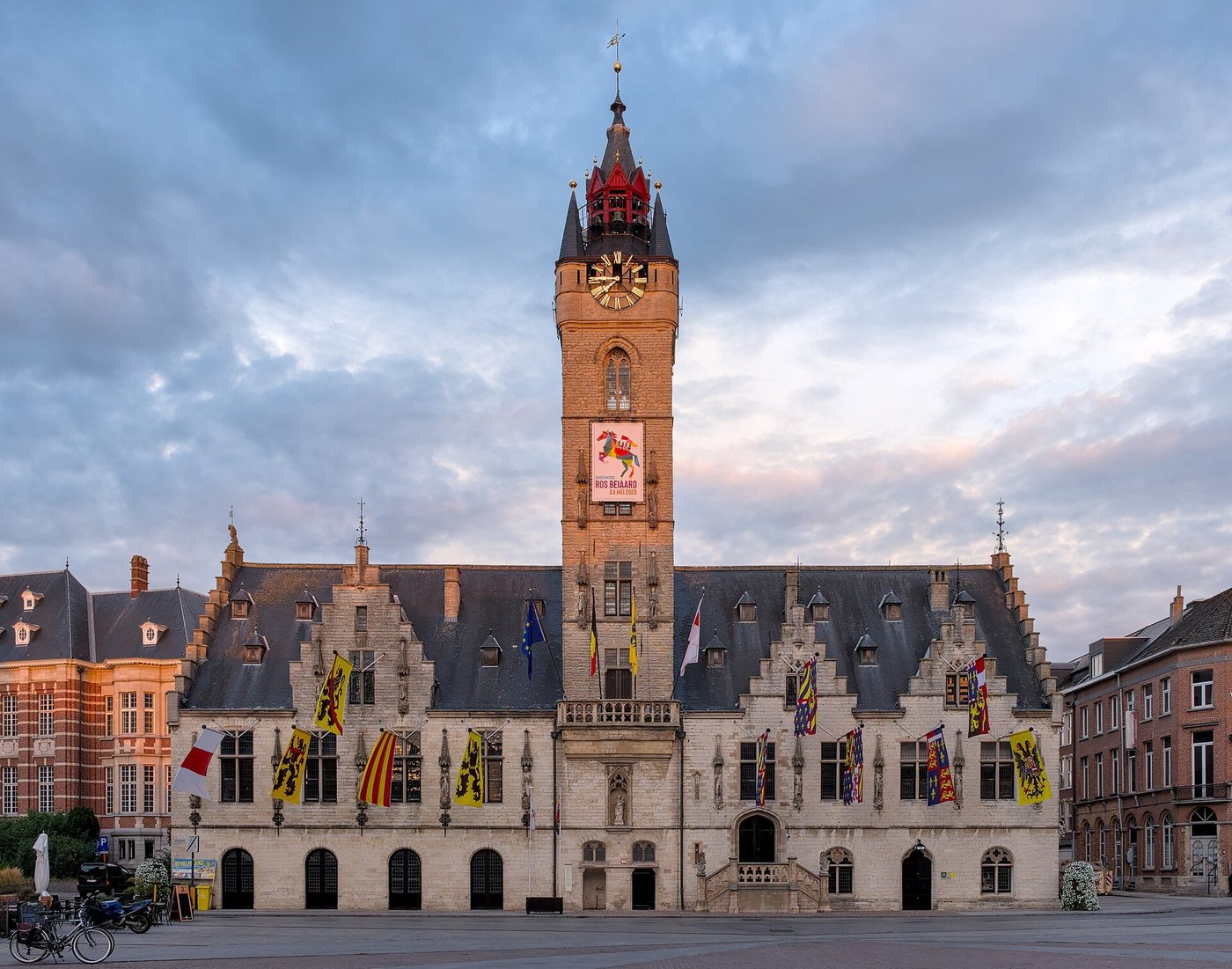 Een stadhuis met een hoge belforttoren in avondlicht, aan een plein.