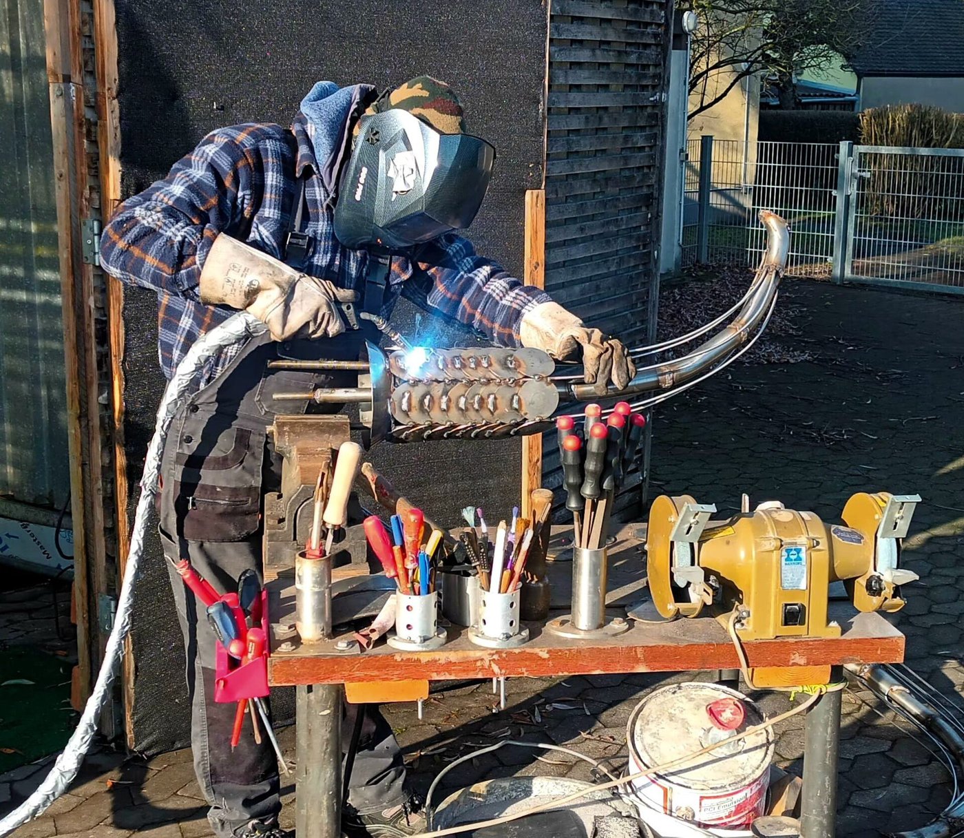 Een lasser met gelaatsscherm aan het werk aan een werkbank buiten, met vonken.