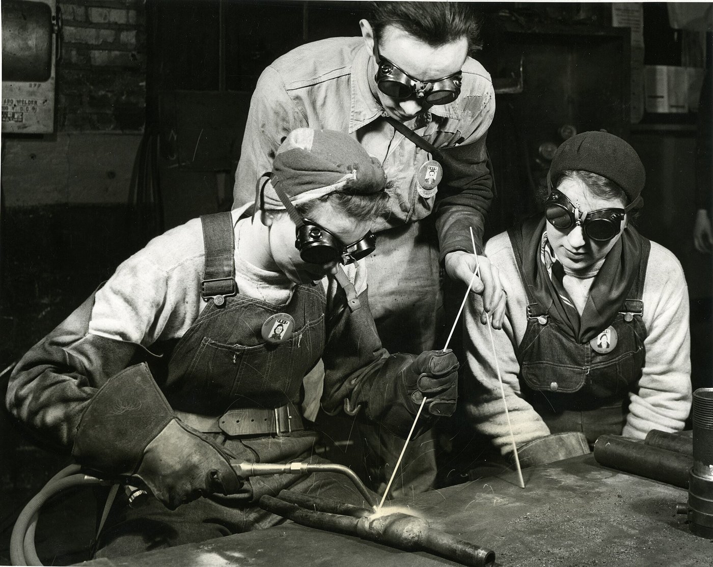 Een historische zwart-witfoto van een leermeester die twee lassers in opleiding aanwijzingen geeft.