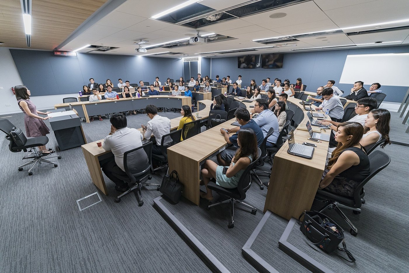 Een collegezaal met cursisten aan lange tafels en een spreker vooraan.