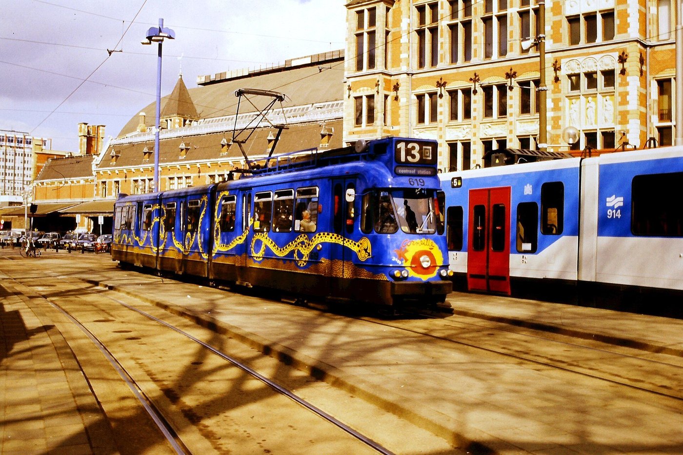 Een blauwe tram op een druk plein voor Amsterdam Centraal, met reizigers op de halte.