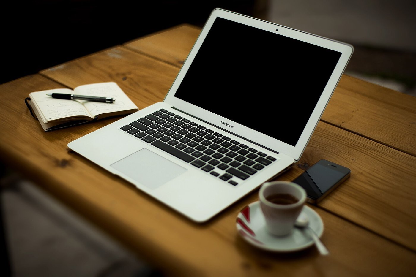 Een laptop op een houten bureau met een notitieboek, een pen, een telefoon en een kop koffie.