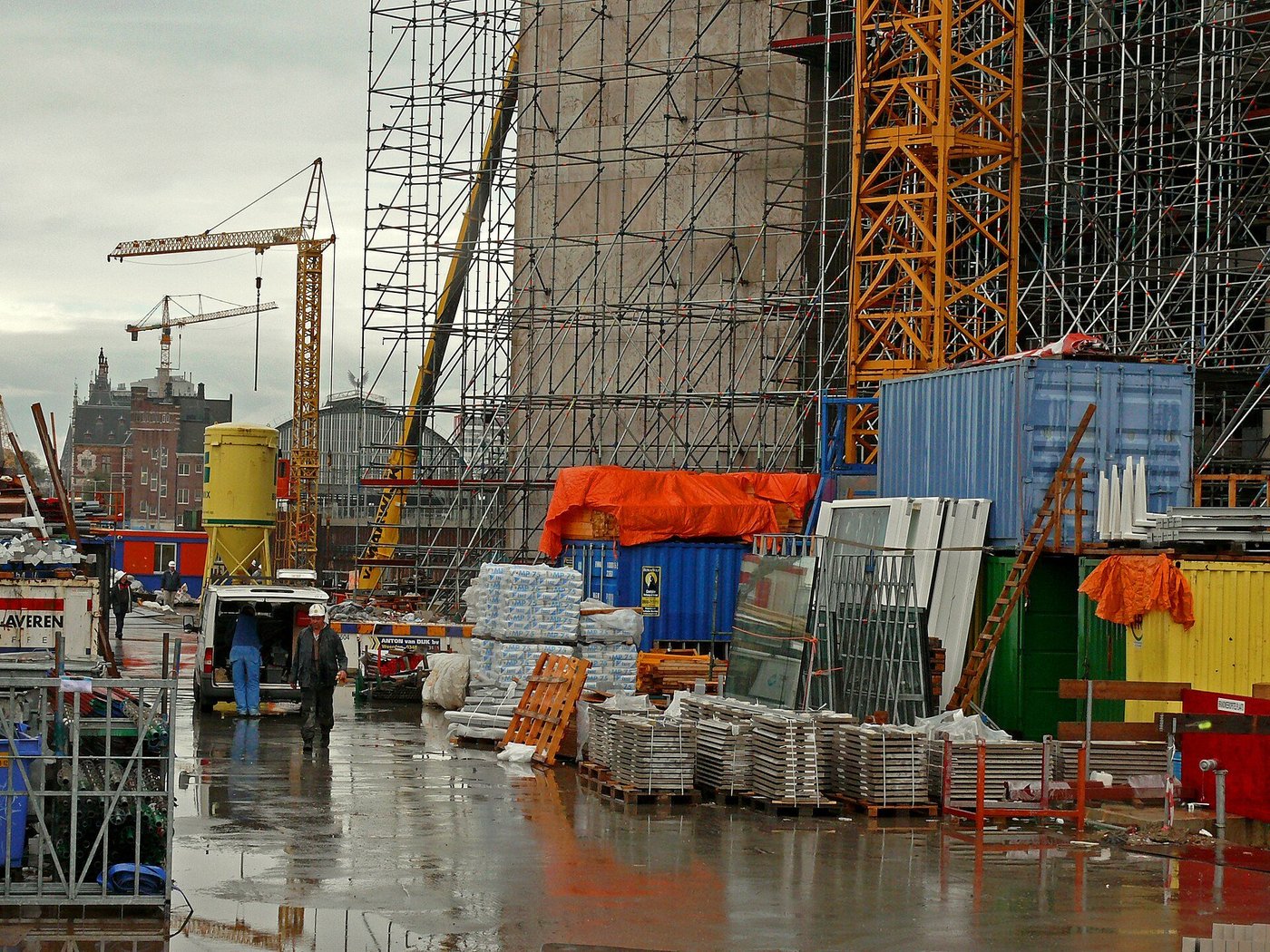 Een bouwplaats in Amsterdam met steigers, containers en kranen aan het water.