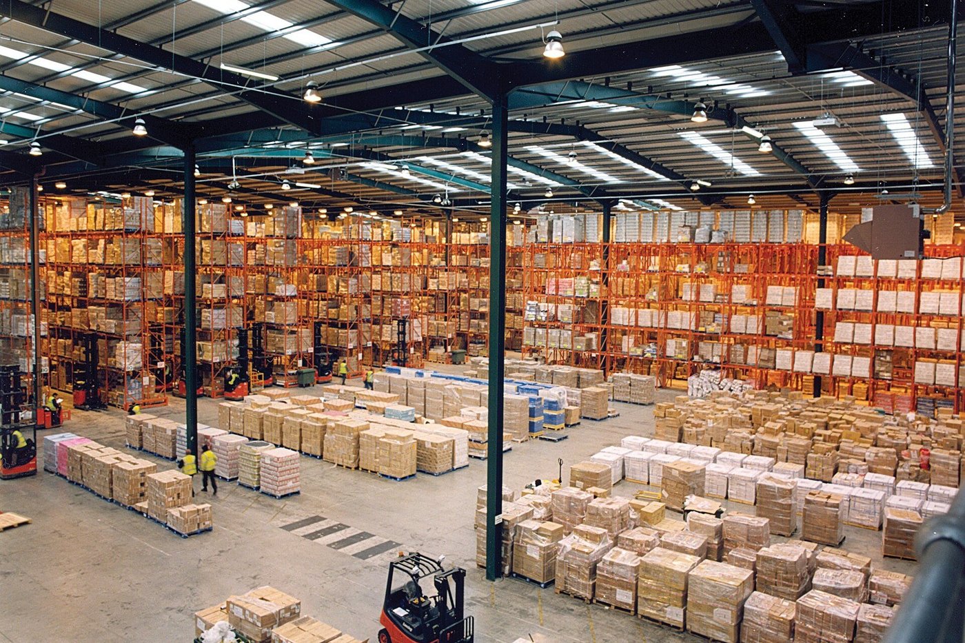 Een grote magazijnhal met pallets op de vloer en een heftruck in de gang.